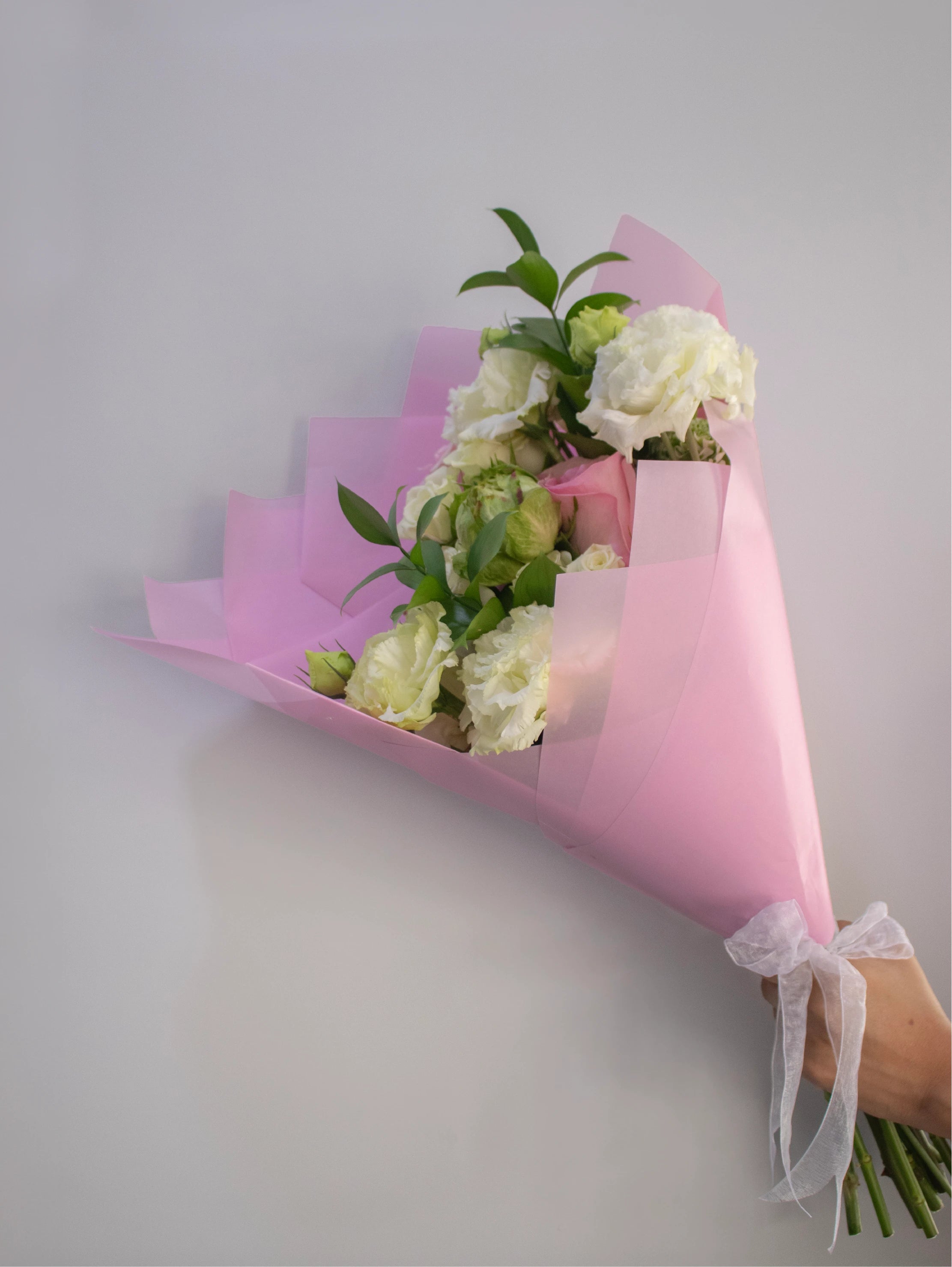 Bouquet of flowers wrapped in pink paper with a white ribbon on a light gray background.