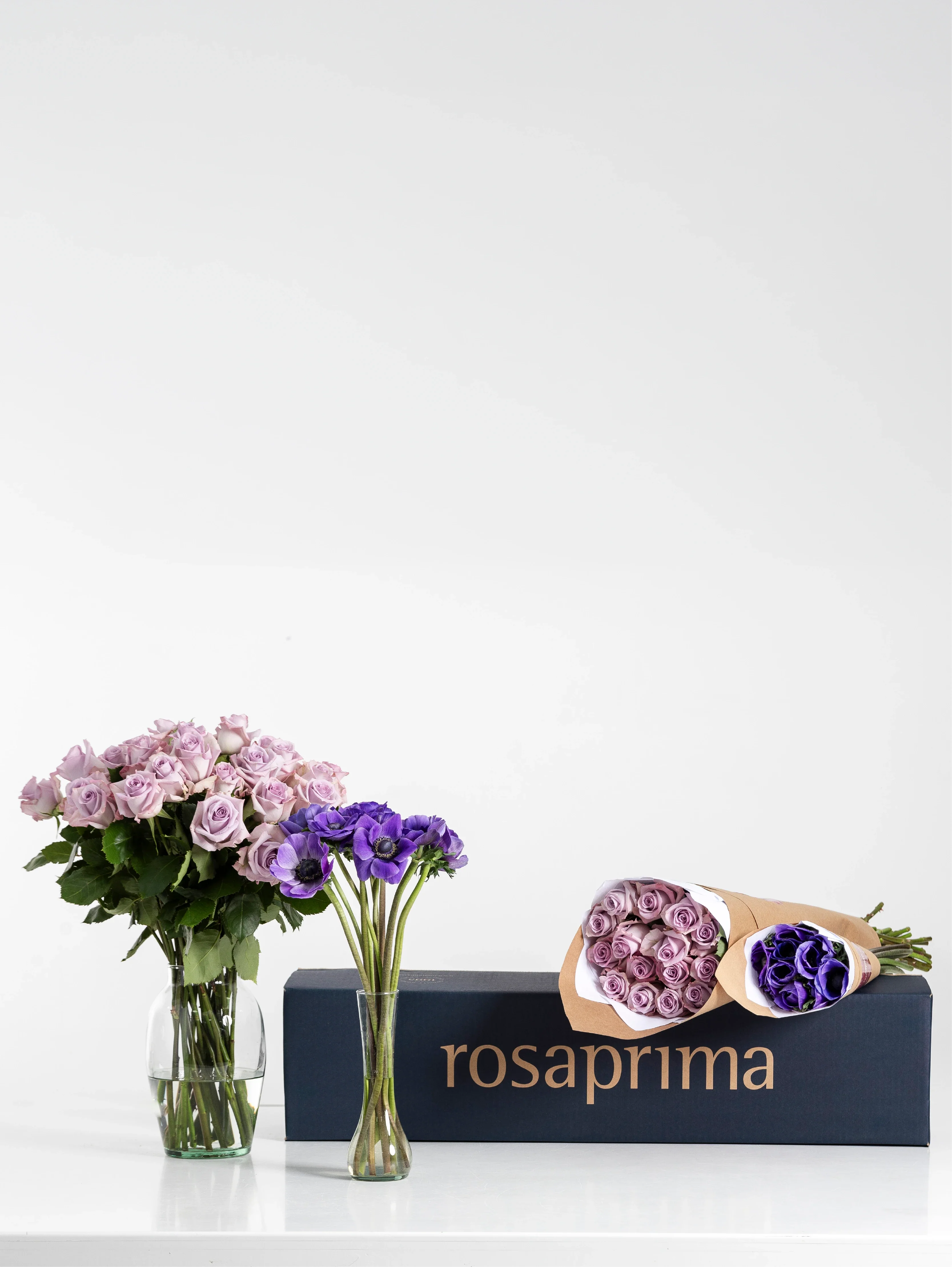 Bouquets of lavender standard roses and lavender mistral anemones with a 'rosaprima' box on a white background.