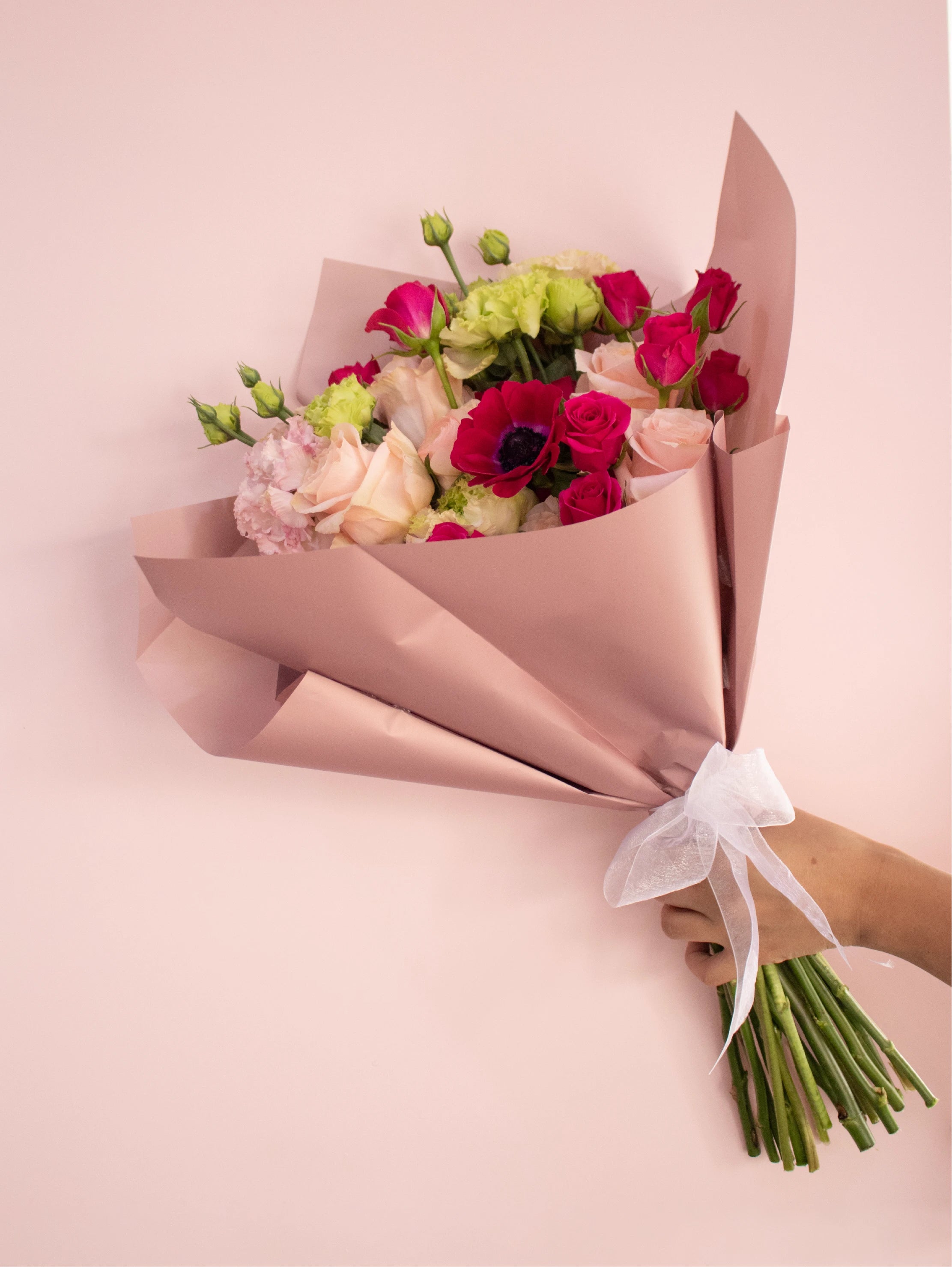 Bouquet of flowers wrapped in brown paper with a white ribbon on a pink background.