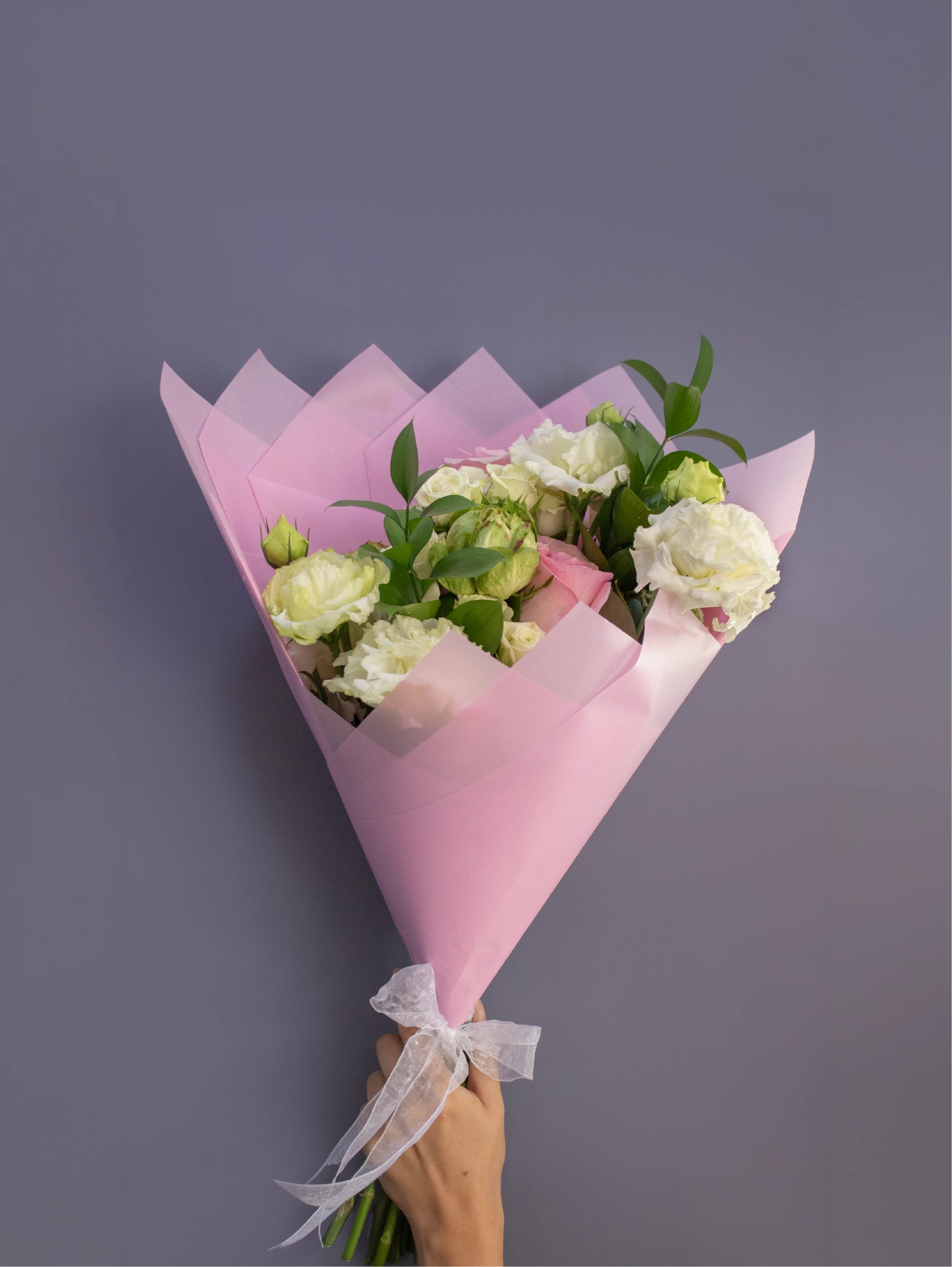 Bouquet of white flowers wrapped in pink paper with a hand holding it against a gray background.